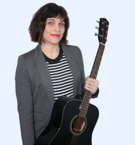 Ali Lipman holding an acoustic guitar while wearing a black and white striped shirt and gray blazer.