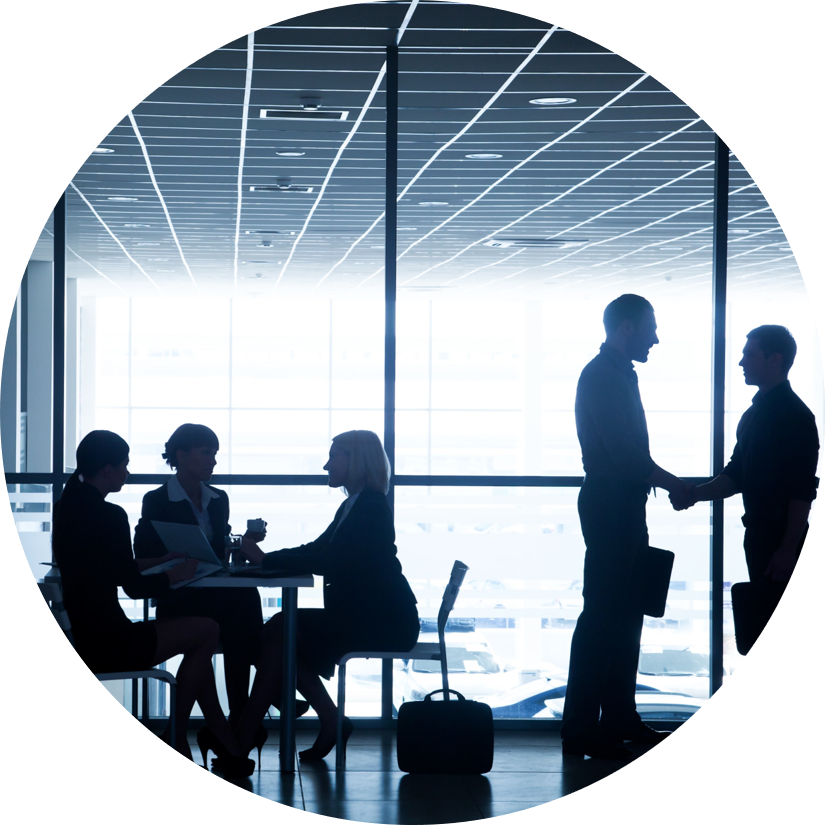 Five colleagues collaborating in an office setting. Three people are sitting at a table in front of a window while two people are shaking hands nearby.