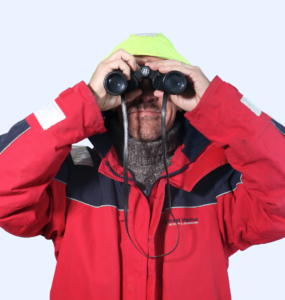 Kevin Cook wearing a red coat and neon yellow beanie while looking through binoculars.