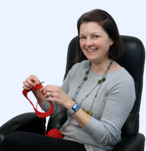 Kim Meyer sitting in a desk chair while knitting with bright red yarn.